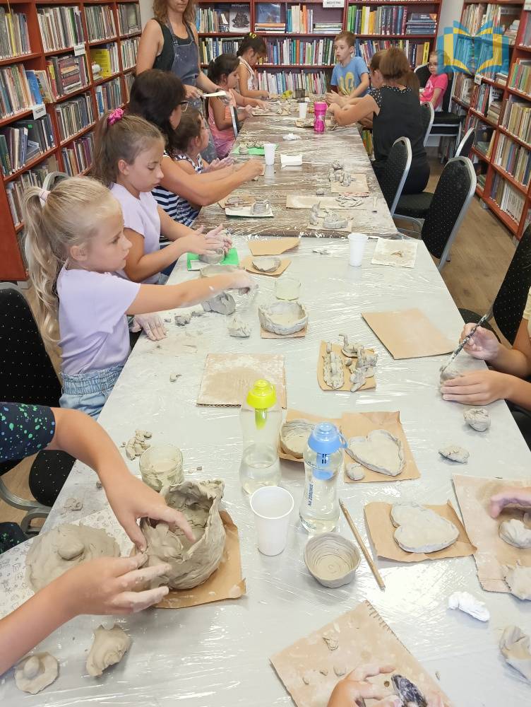 Zdjęcie: Warsztaty ceramiczne w bibliotece. Dzieci siedzą przy długim stole i wykonują różne przedmioty z gliny.